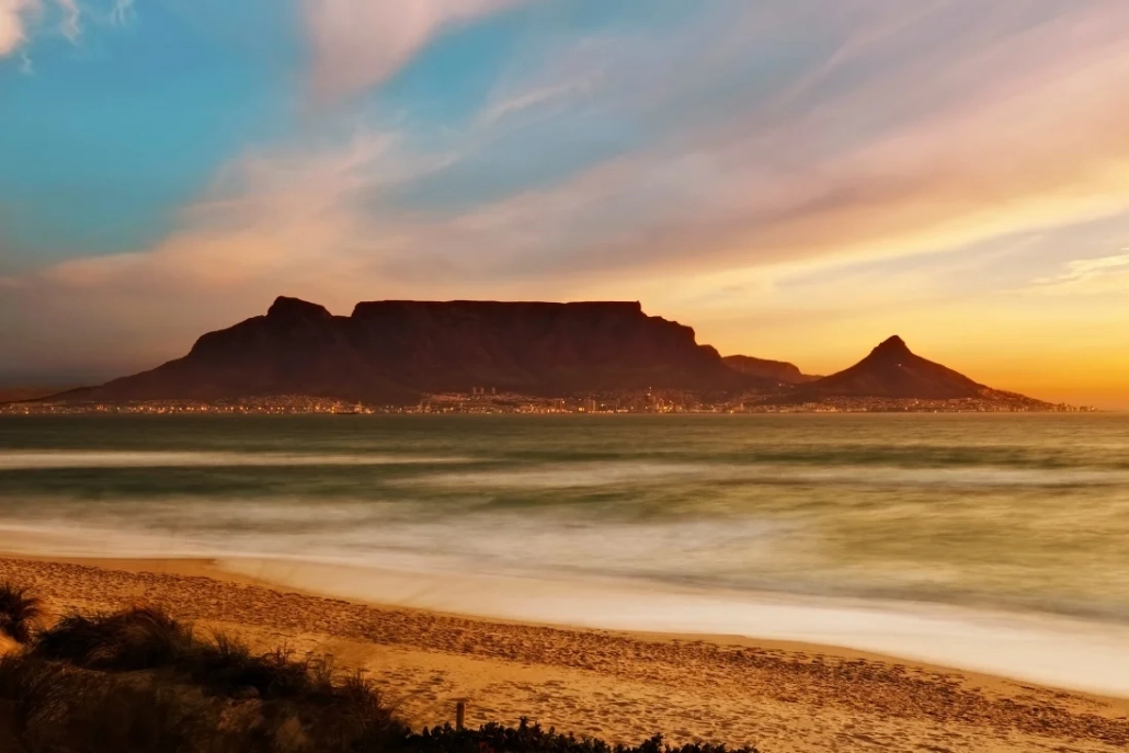 The iconic Table Mountain silhouetted against a vibrant sunset sky, with the city lights reflecting on the ocean