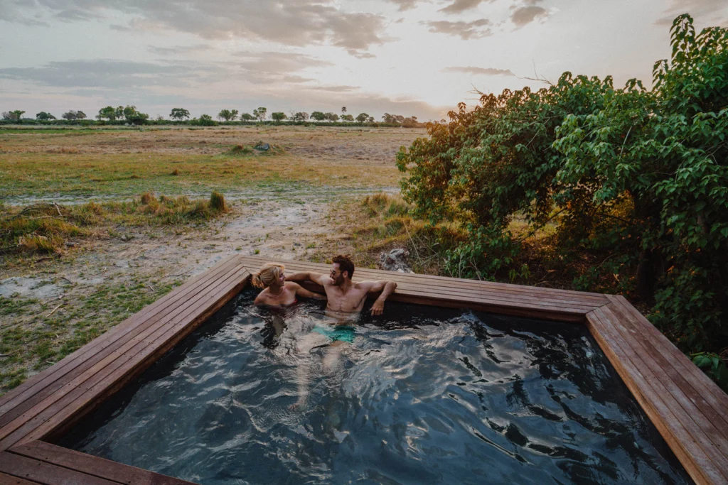 Two people sit in a square plunge pool surrounded by the African wilderness