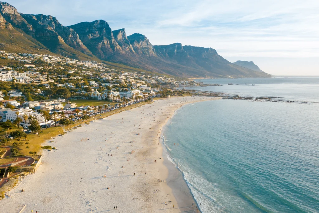 A scenic view of Camps Bay Beach with white sand, turquoise waves, and the Twelve Apostles mountain range in the background.