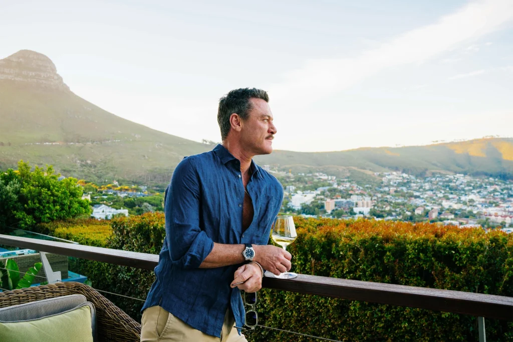 Actor Luke Evans enjoying a glass of wine on the balcony at Atzaró Cape Town, with views of the city and Lion’s Head in the background.