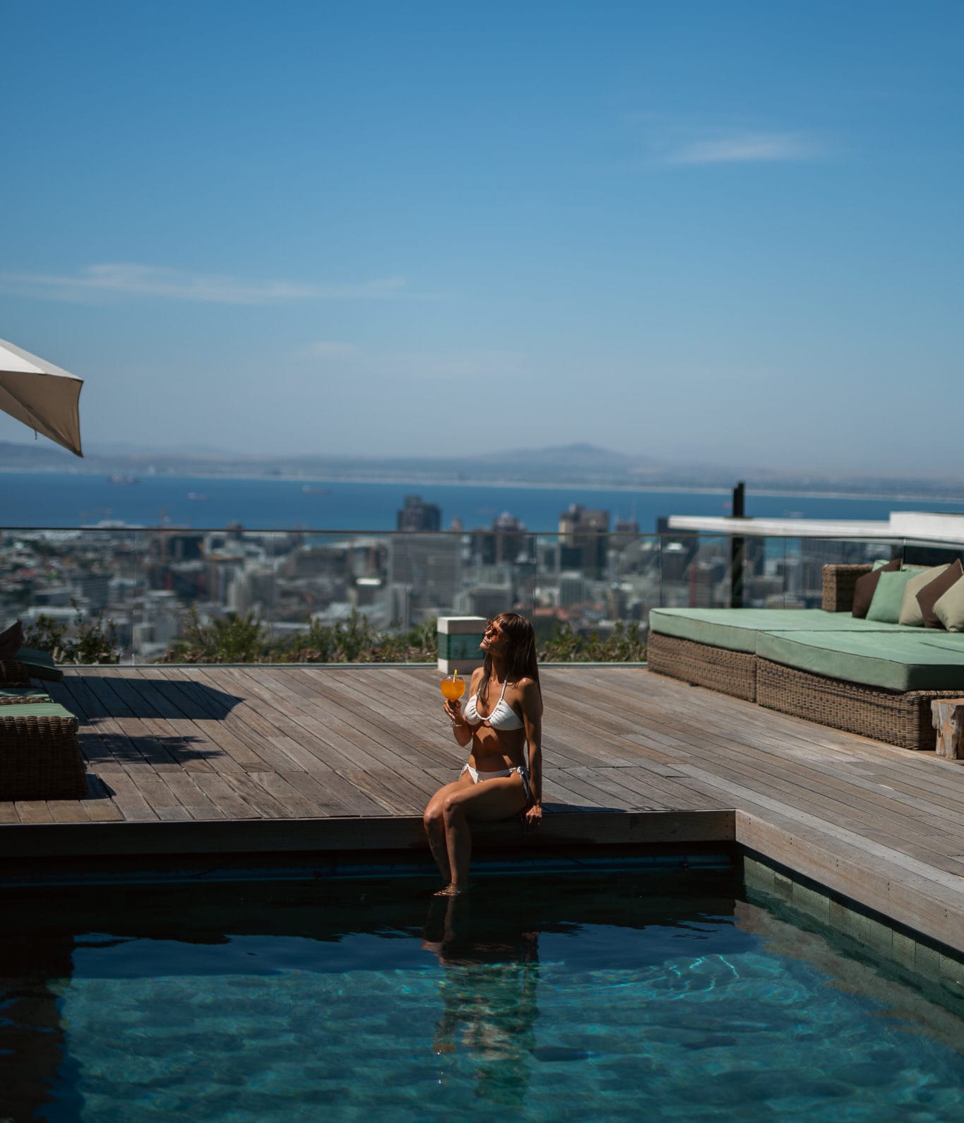 A woman relaxing by a rooftop swimming pool on a wooden deck at Atzaró Cape Town, holding a drink with panoramic views of the city and the Atlantic Ocean.