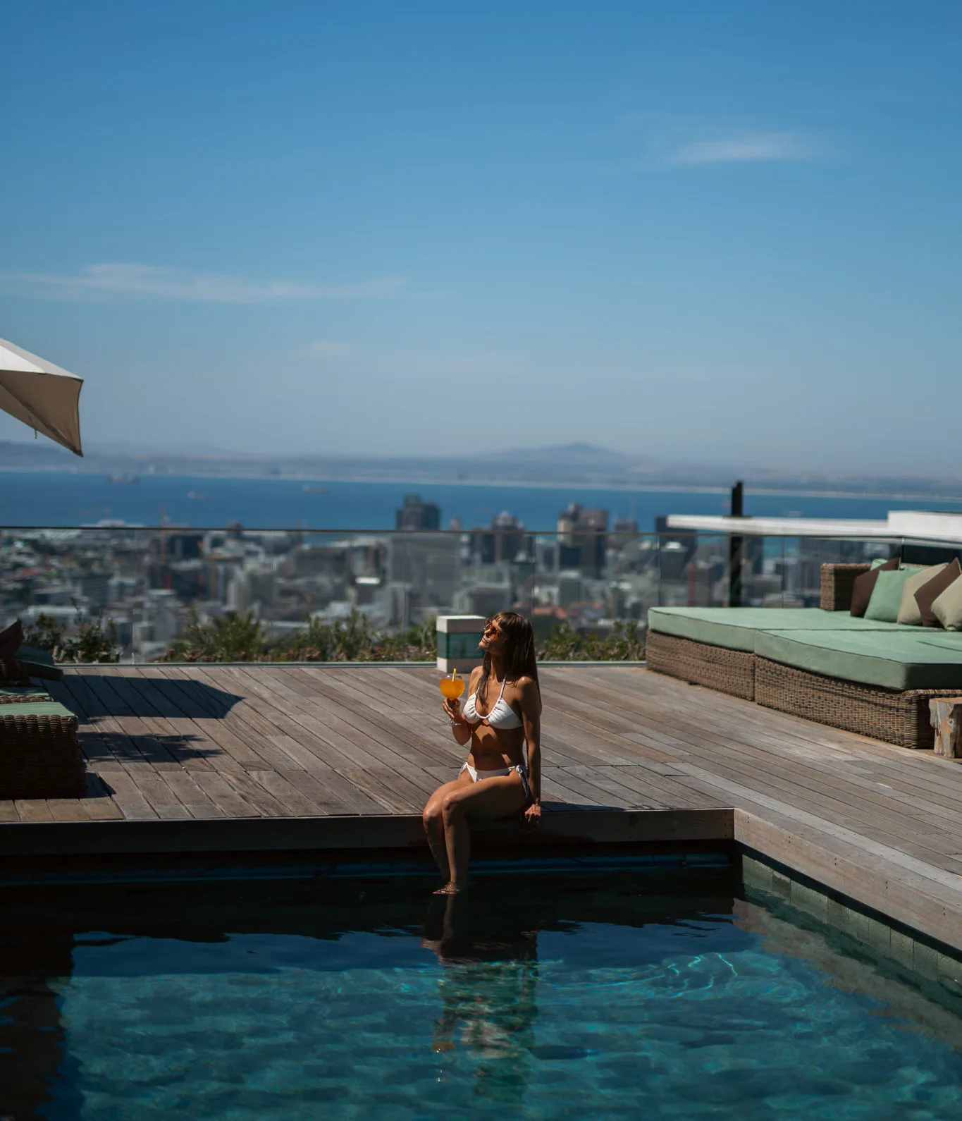 A woman relaxing by a rooftop swimming pool on a wooden deck at Atzaró Cape Town, holding a drink with panoramic views of the city and the Atlantic Ocean.