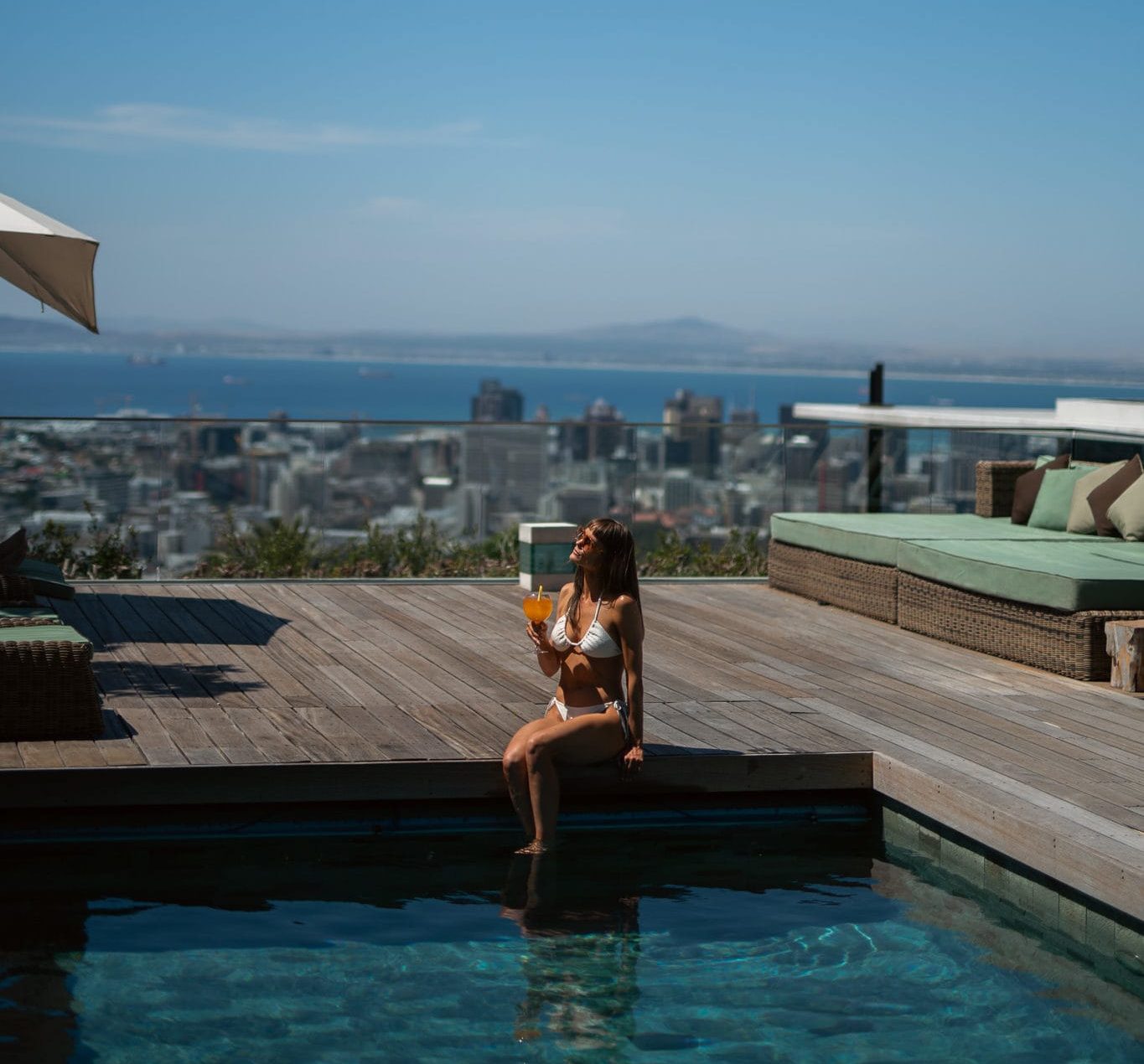 A woman relaxing by a rooftop swimming pool on a wooden deck at Atzaró Cape Town, holding a drink with panoramic views of the city and the Atlantic Ocean.