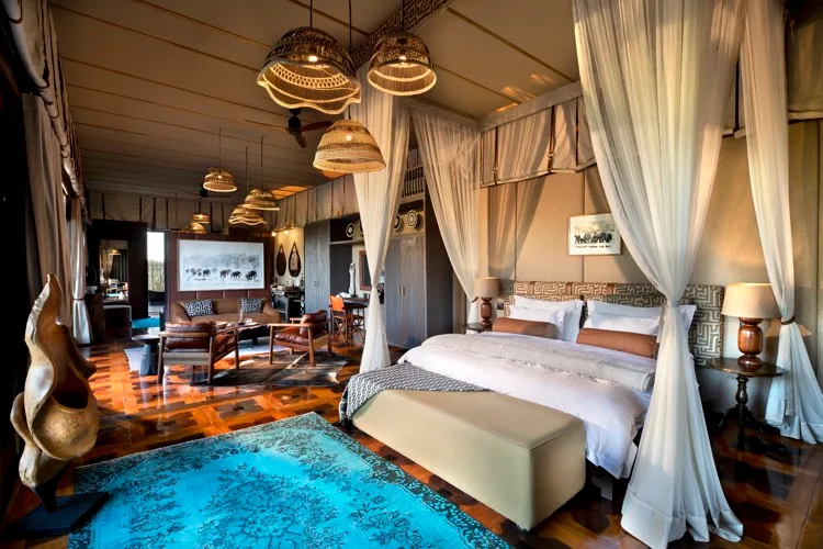 Luxurious interior of an Atzaro Okavango Camp suite featuring a canopy bed with sheer white drapes, woven pendant lights, polished parquet flooring, and an elegant lounge area.