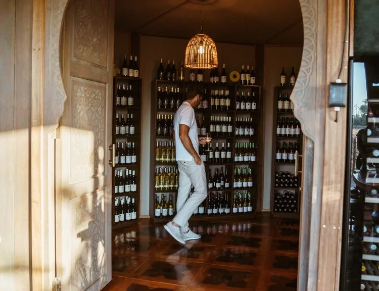 A guest holding a wine glass while browsing the extensive wooden bottle racks inside the wine cellar at Atzaro Okavango Camp, viewed through ornate carved doors.