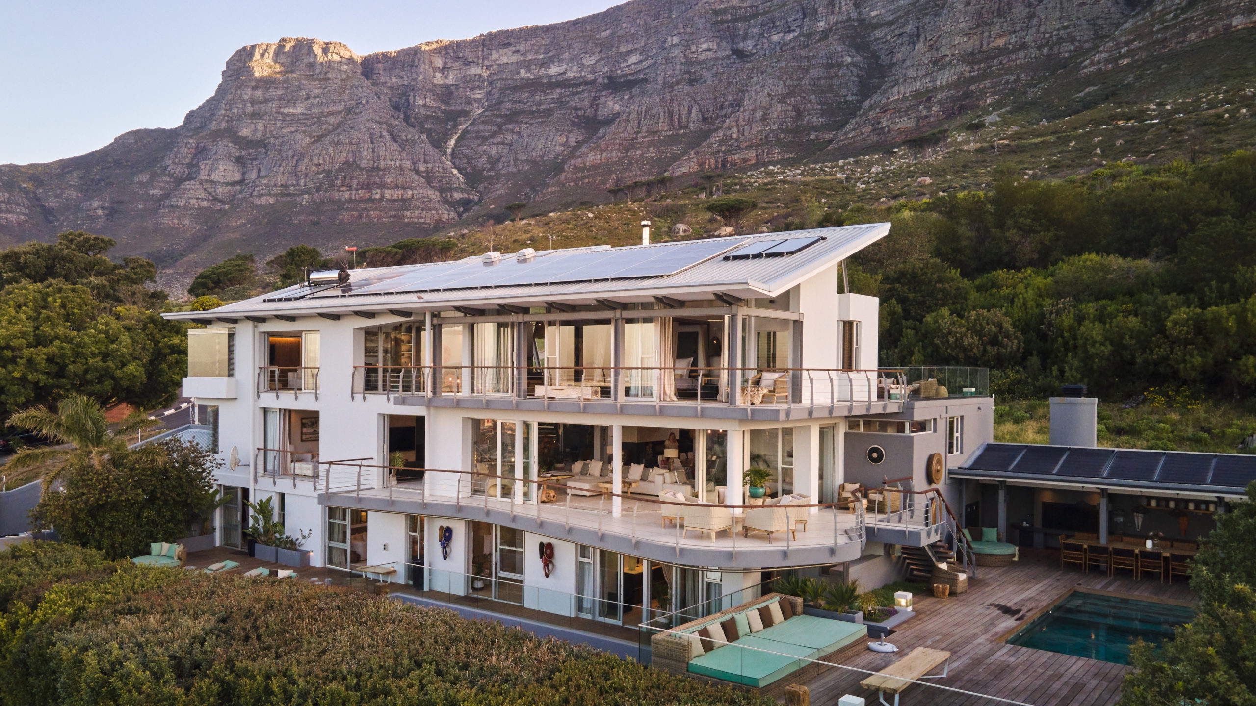Aerial view of Atzaró Cape Town: a white three-level hotel with solar panels, glass terraces and a turquoise pool, set against the brown cliffs of Table Mountain and green hillside at the foot of the mountain.