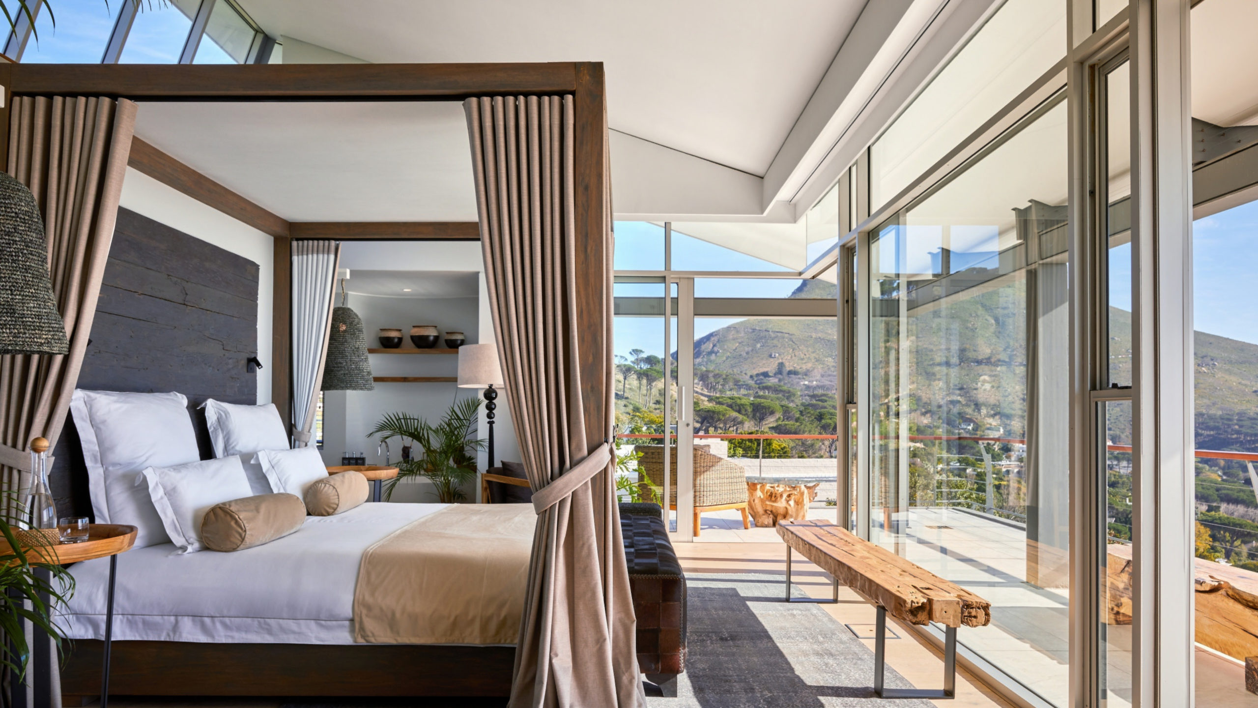 Suite at Atzaró Cape Town with a wooden four-poster bed, white pillows, a rustic wood headboard and linen curtains, under a high glass ceiling that frames the rocky cliffs of Table Mountain above.