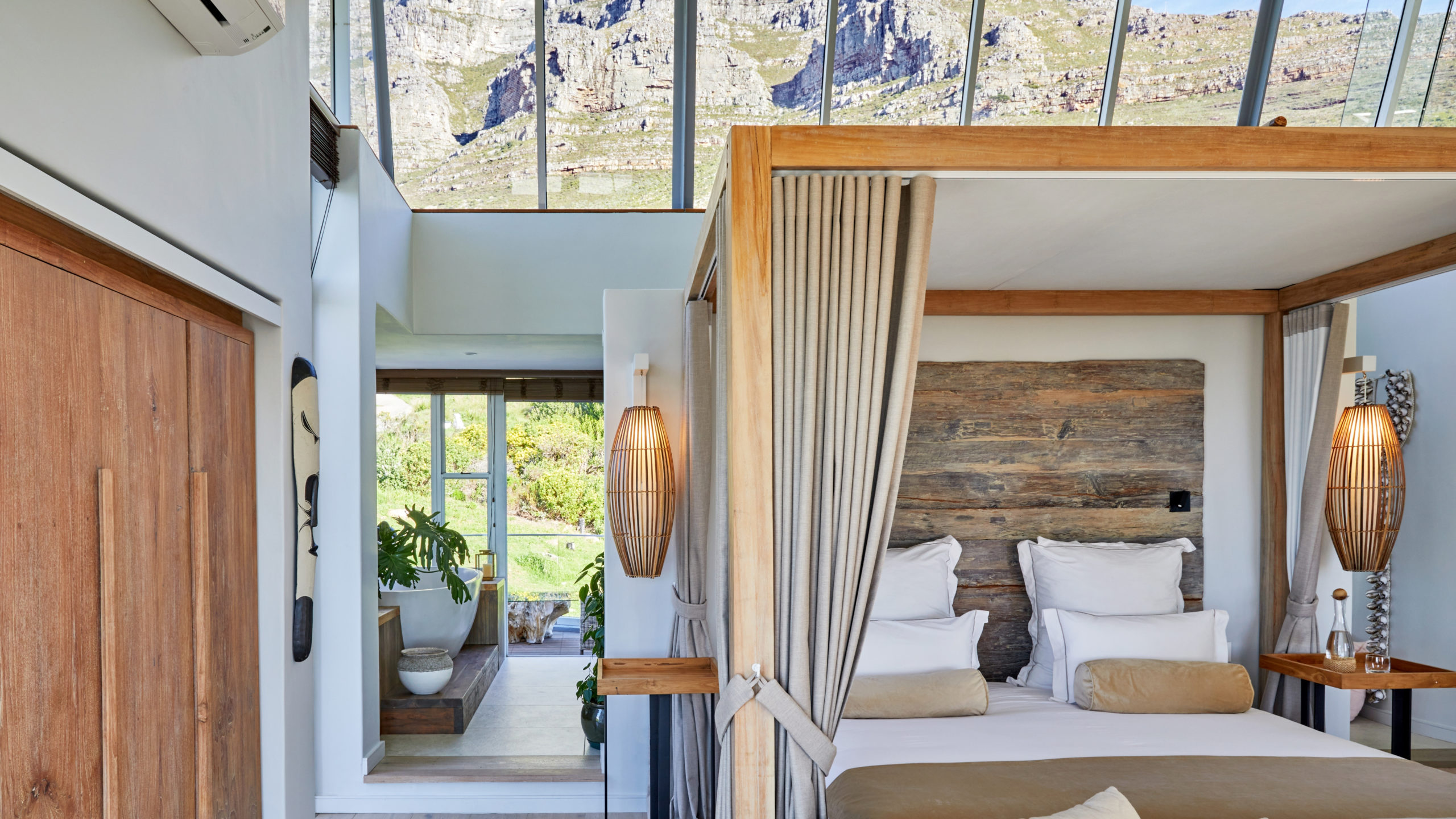 Bright suite bedroom with a wooden canopy bed, cream and beige linens, tall linen curtains, a potted palm and a dark gray rug, beside floor-to-ceiling glass doors that open to a balcony with hill views.