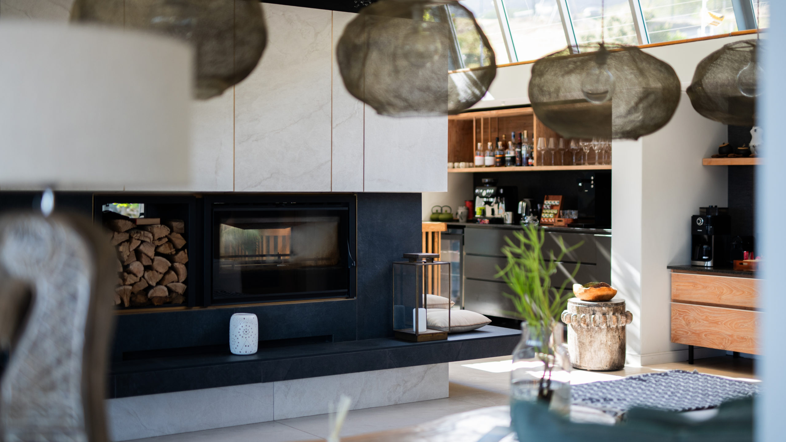 Cozy lounge at Atzaró Cape Town with a black fireplace, a neat stack of firewood, woven mesh pendant lamps, pale marble walls and a wooden bar shelf with bottles, a warm place to end a winter evening on a Cape Town honeymoon.