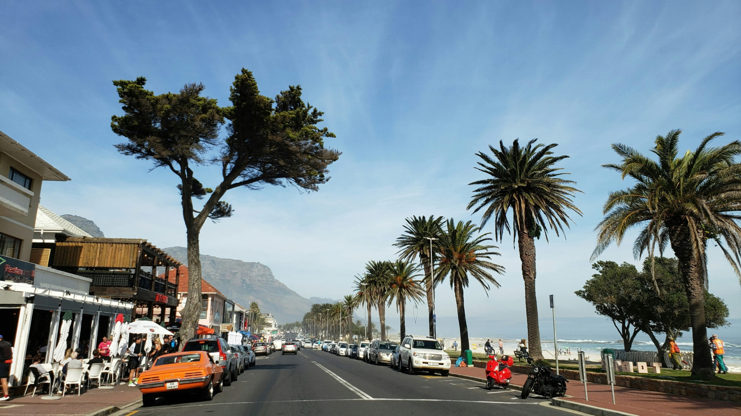 Parked cars along a Camps Bay street with restaurants, the beach and the mountains in the background