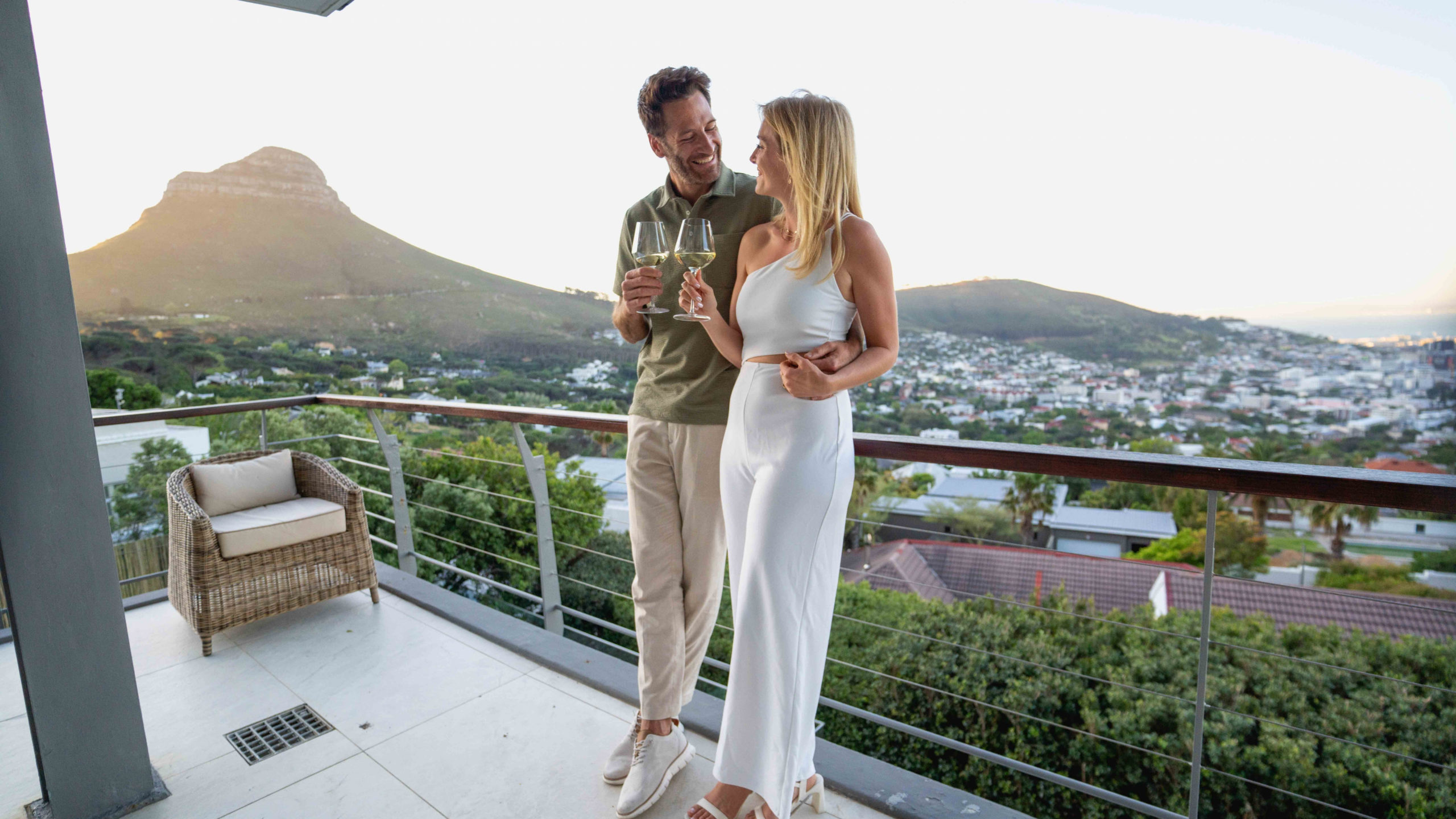 Couple in olive and white outfits toasting with wine glasses on a terrace at golden hour, with Lion’s Head and Cape Town rooftops glowing softly behind them, a romantic sunset moment in Cape Town.