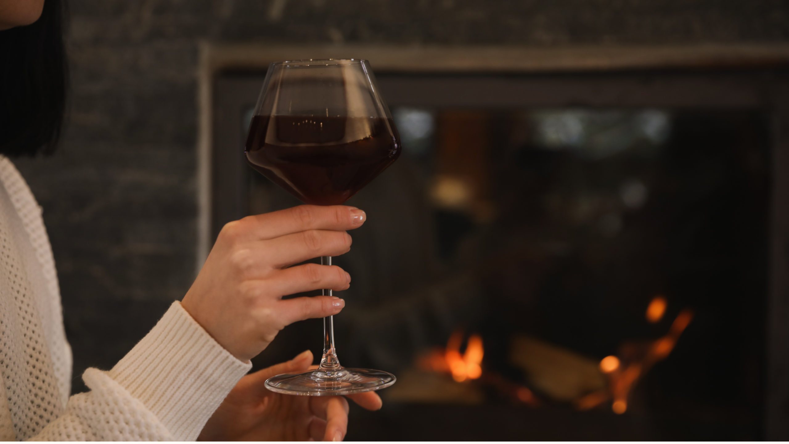 Close-up of a hand holding a large glass of deep red wine in front of a glowing fireplace, with orange flames in the dark background and a white knit sleeve in view, a cozy winter wine moment for couples in Cape Town.