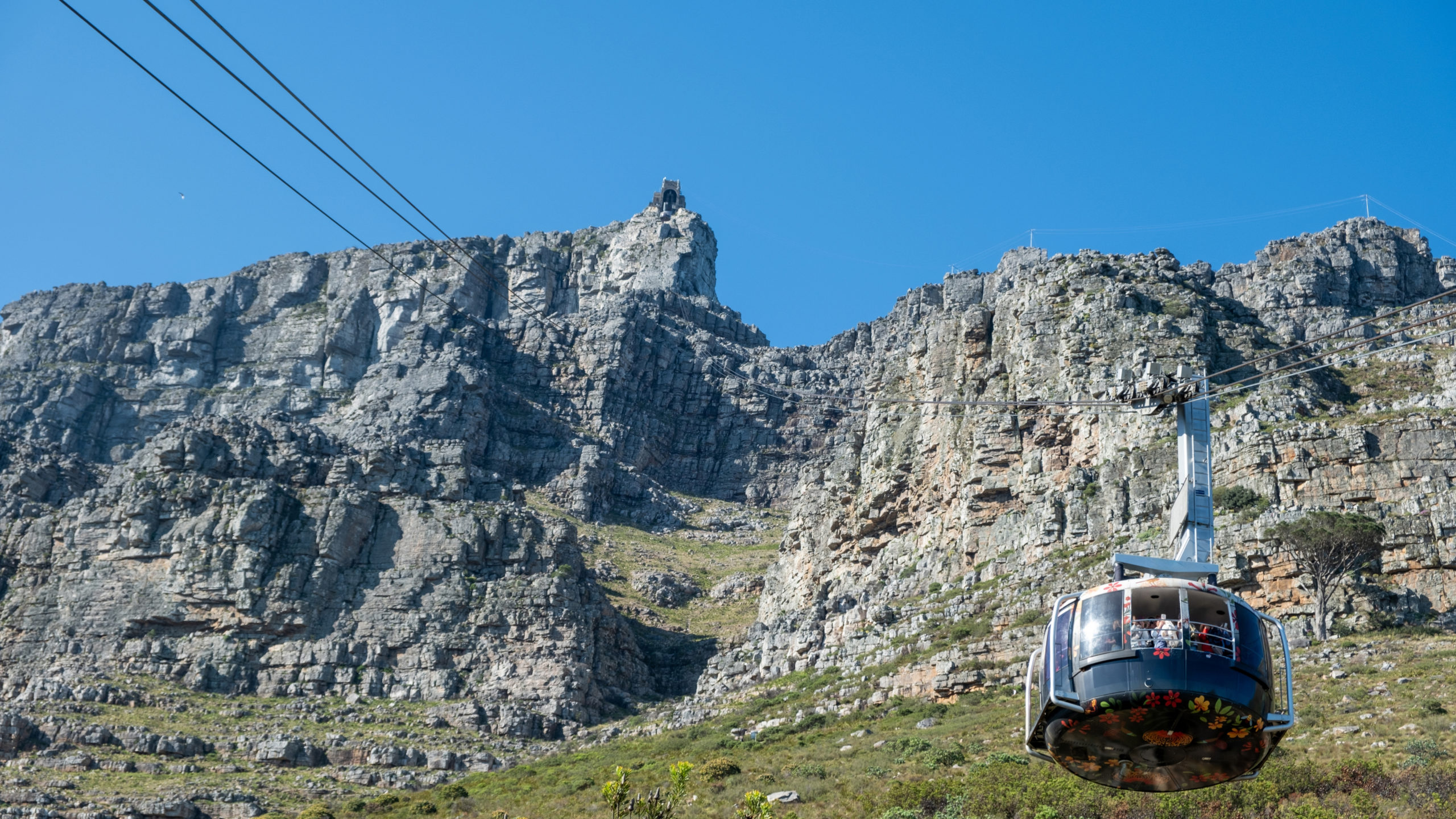 Table Mountain cable car, a dark blue cabin, climbing the gray rocky cliffs toward the upper station beneath steel cables and a bright cloudless blue sky, the ride that leads to the best sunset views.